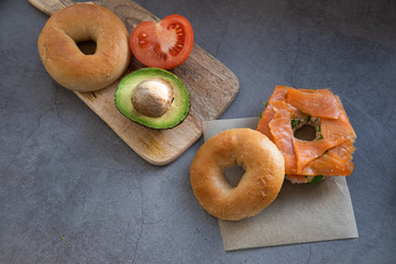 Salmon bagel on baking paper and wooden table with tomato, avocado and a bagel.