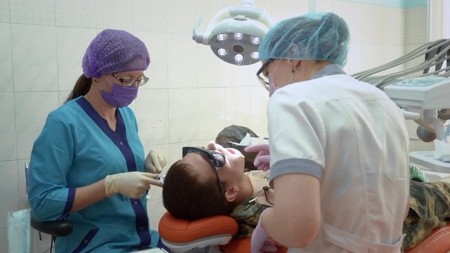 Patient at the dentist's appointment. The dentist and her assistant are treating the patient in military uniform. The dentist fills the tooth.