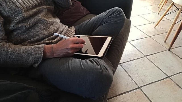 Uomo in abbigliamento casual che disegna al tablet a casa seduto a gambe incrociate sul divano