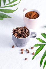 Ceramic cups with freshly roasted coffee beans and fresh ground aromatic coffee on a light background and green palm leaves.
