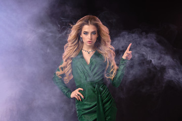 Blonde lady in green stylish dress and jewelry. Pointing at something by forefinger, posing on black smoky background. Close up, copy space