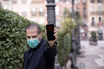 uomo rasato in giacca indossa una mascherina di protezione e  ci mostra lo schermo di un cellulare con su scritto: Stop Covid-19 