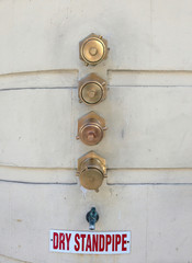 Shiny brass water outlets for the fire department on an outside building wall labeled DRY STANDPIPE