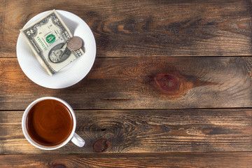 White cup with invigorating espresso coffee and saucer with two American dollar bill and coin. On dark wooden background. Payment or tip. Top view. Copy space for text.