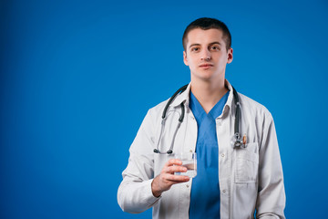 Young male doctor in medical uniform with stethoscope holding glass of clear water isolated on blue