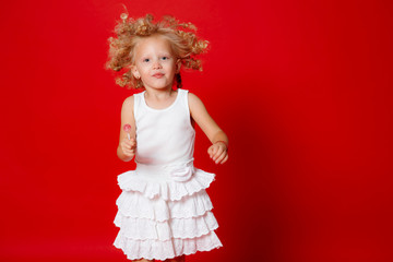 Beautiful little curly blonde girl in a white dress eating lollipop jumping isolated on red