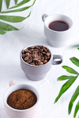 Ceramic cups with freshly roasted coffee beans and fresh ground aromatic coffee on a light background and green palm leaves.
