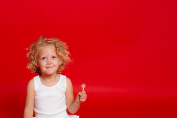 Beautiful little curly blonde girl eating lollipop sitting on the floor isolated on red