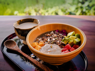 vegan fruit smoothie bowl with wooden spoon, granola, chia, seeds, mango, dragon fruit, watermelon, avocado and a cup of almond milk on a tray for healthy breakfast with green grass in background