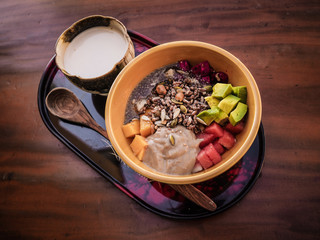 birds eye view of a vegan fruit smoothie bowl with wooden spoon, granola, nuts, chia, seeds, mango, dragon fruit, watermelon, avocado and a cup of almond milk on a tay on a table for healthy breakfast