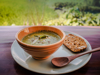 close-up of a healthy vegan green soup with pumpkin and sunflower seeds, salad, bread and wooden spoon in a bowl on a plate on table with rice field in the beackround