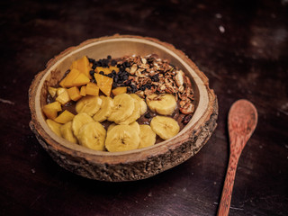 close-up of a vegan fruit smoothie bowl with wooden spoon, granola, nuts, banana mango and cocoa on a table for a healthy breakfast 
