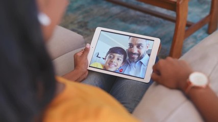 Rear view of young woman relaxing on couch while talking to her husband and son using digital tablet at home. Mature middle eastern man and child communicate through video chat on laptop. - Powered by Adobe