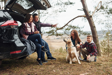 Happy family sitting and having fun with their dog near modern car outdoors in forest