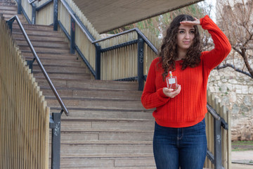 Portrait of attractive caucasian young woman model with cologne perfume bottle peeks out someone, keeps hand near forehead, in the park, orange sweater and jeans. Place for your text in copy space.