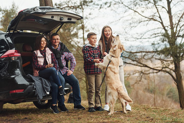 Happy family have fun with their dog near modern car outdoors in forest