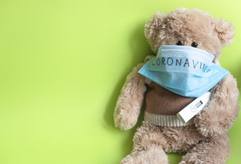 Soft toy bear in a protective medical mask with the inscription "coronavirus" and a thermometer, sitting on a green background. The concept of home education from the COVID-19 quarantine
