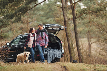 Happy mature couple have a walk with their dog in autumn or spring forest near modern car