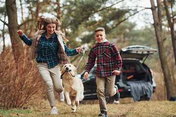 Cheerful girl with brother have walk with their dog outdoors in forest at autumn or spring season near car