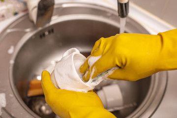Close up image of hands in yellow protective rubber gloves washing white cup under running tap water.