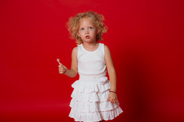 Confused blonde curly girl in white dress holding lollipop isolated on red