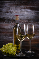 White wine bottle and two glasses with grapes on a table on wooden background