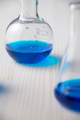 Close-up of two glass test tubes with blue liquid, the first out of focus, on white wooden table, vertical