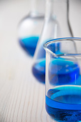 Close-up of three glass test tubes with blue liquid and selective focus, on defocused white wooden table, vertical