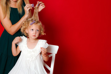 Mother curls daughter hair. Little blonde girl sitting on a white chair eating lollipop isolated on red