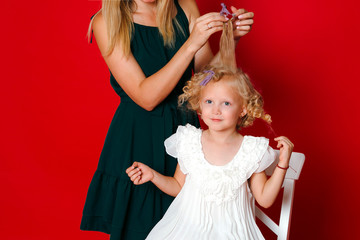 Mother curls daughter hair. Little blonde girl sitting on a white chair eating lollipop isolated on red