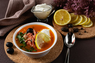 Soup Solyanka is a combined meat soup with smoked meat , various types of meat and tomato paste, serving dishes with sour cream, lemon and fresh herbs. Black olives on the table and a sprig of fresh f