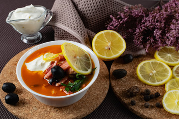 Soup Solyanka is a combined meat soup with smoked meat , various types of meat and tomato paste, serving dishes with sour cream, lemon and fresh herbs. Black olives on the table and a sprig of fresh f