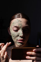 Beautiful Young Woman with organic cosmetic mask (scrub) looking her smartphone. Facial treatment . Cosmetology , beauty and spa