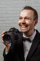 Young handsome male photographer with a beautiful smile in a black jacket, white shirt and black bow-tie holds a camera in his hand and smiles.