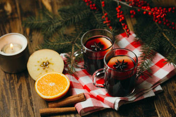 Two glass cups with mulled wine with citrus, apple and spices on checked tablecloth. Winter holiday mood with fur tree branches and red berries.
