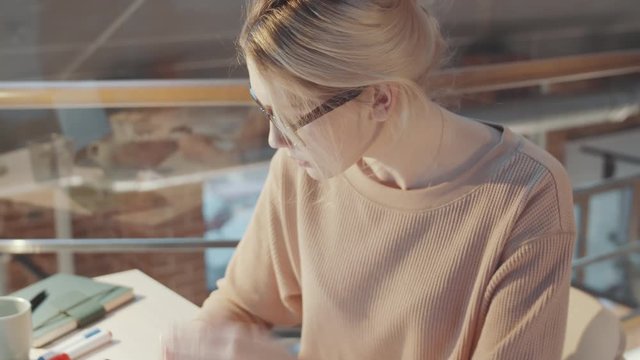 Tilt down shot of blonde female designer in glasses holding felt tip pen and checking fashion illustration while working at desk in studio