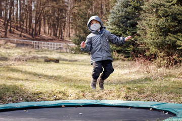Small boy jumping on a trampoline during a pandemic, wearing fabric facemask and hoodie, looking...