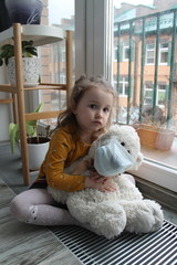 A little girl and a toy bear in a medical mask near the window, the need to stay at home because of the epidemic. Schools and kindergartens are under quarantine.