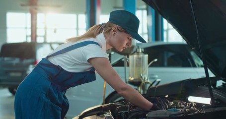 Female Caucasian young auto mechanic standing at open car and repairing motor in garage, male colleague coming to help. Woman and man working in automobile repairment salon indoors. Service concept.