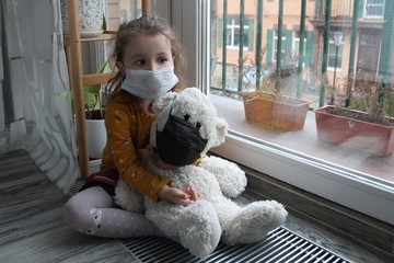 A girl and a toy bear in medical masks near the window, looking at the street. Schools and kindergartens are under quarantine. Concept of isolation and quarantine in the context of a pandemic. 