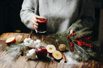 Hot mulled wine in the woman hands in grey sweater. Beautiful Christmas decorations lying on the table: fur-tree branches, red berries, apples, oranges, cotton flower. Festive mood.