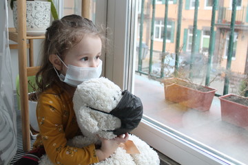 A girl and a toy bear in medical masks near the window, looking at the street. Schools and kindergartens are under quarantine. Concept of isolation and quarantine in the context of a pandemic. 