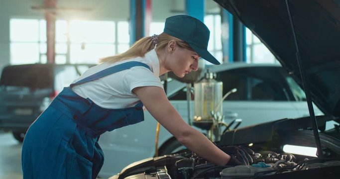 Female Caucasian young auto mechanic standing at open car and repairing motor in garage, male colleague coming to help. Woman and man working in automobile repairment salon indoors. Service concept.