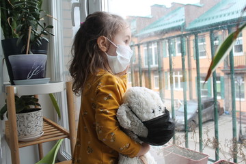 A girl and a toy bear in medical masks near the window , looking at the street.Schools and kindergartens are under quarantine. Concept of isolation and quarantine in the context of a pandemic. 