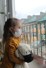 A girl and a toy bear in medical masks near the window , looking at the street.Schools and kindergartens are under quarantine. Concept of isolation and quarantine in the context of a pandemic. 