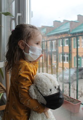 A girl and a toy bear in medical masks near the window , looking at the street.Schools and kindergartens are under quarantine. Concept of isolation and quarantine in the context of a pandemic. 