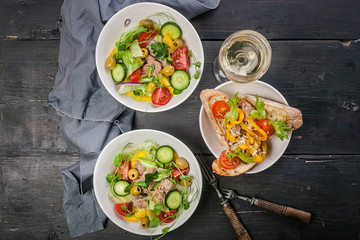 Top view white wine in a glass, salads with vegetables and micro-greens in white bowls, canapes with pate and sweet pepper on a dark wooden table.