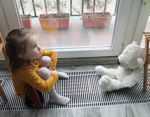 A little girl and a toy bear sit near the window, having to stay at home because of the epidemic. Concept of isolation and quarantine in the context of a pandemic. 2019 coronavirus-nCoV.