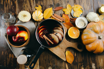 Mulled wine hot drink with citrus, apple and spices in aluminum pot on wooden background. Autumn mood with pumpkins, candles and leaves.