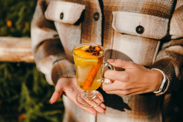 Woman in checked coat holding glass cup of delicious white mulled wine with a cinnamon stick in her...